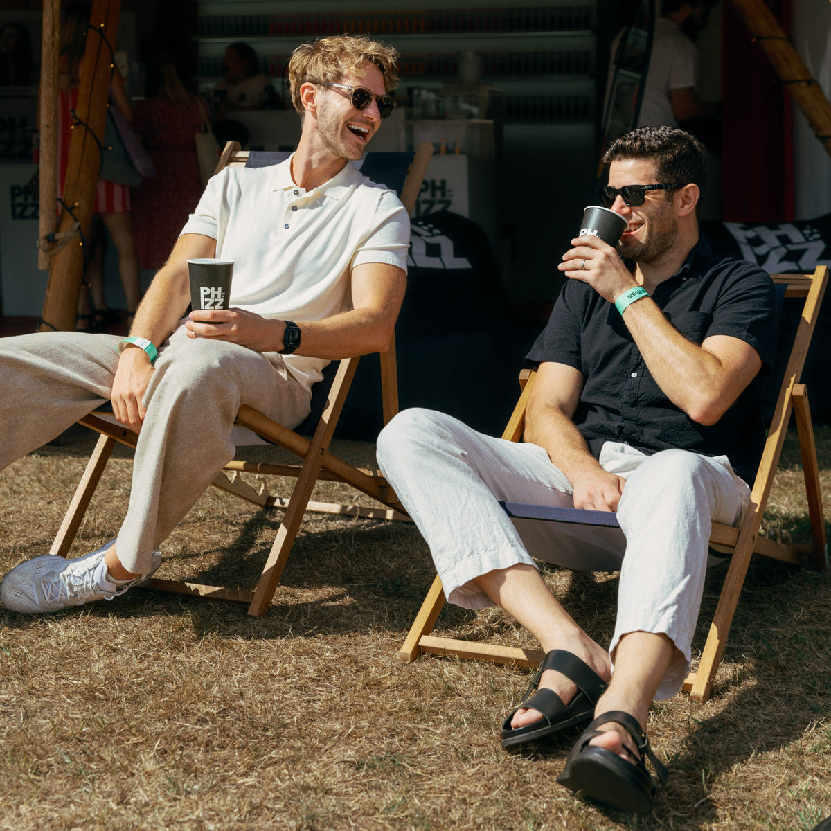 Two men sitting in wooden folding chairs under a beige tent at an outdoor festival, with a 'Hydration Station' sign visible behind them.