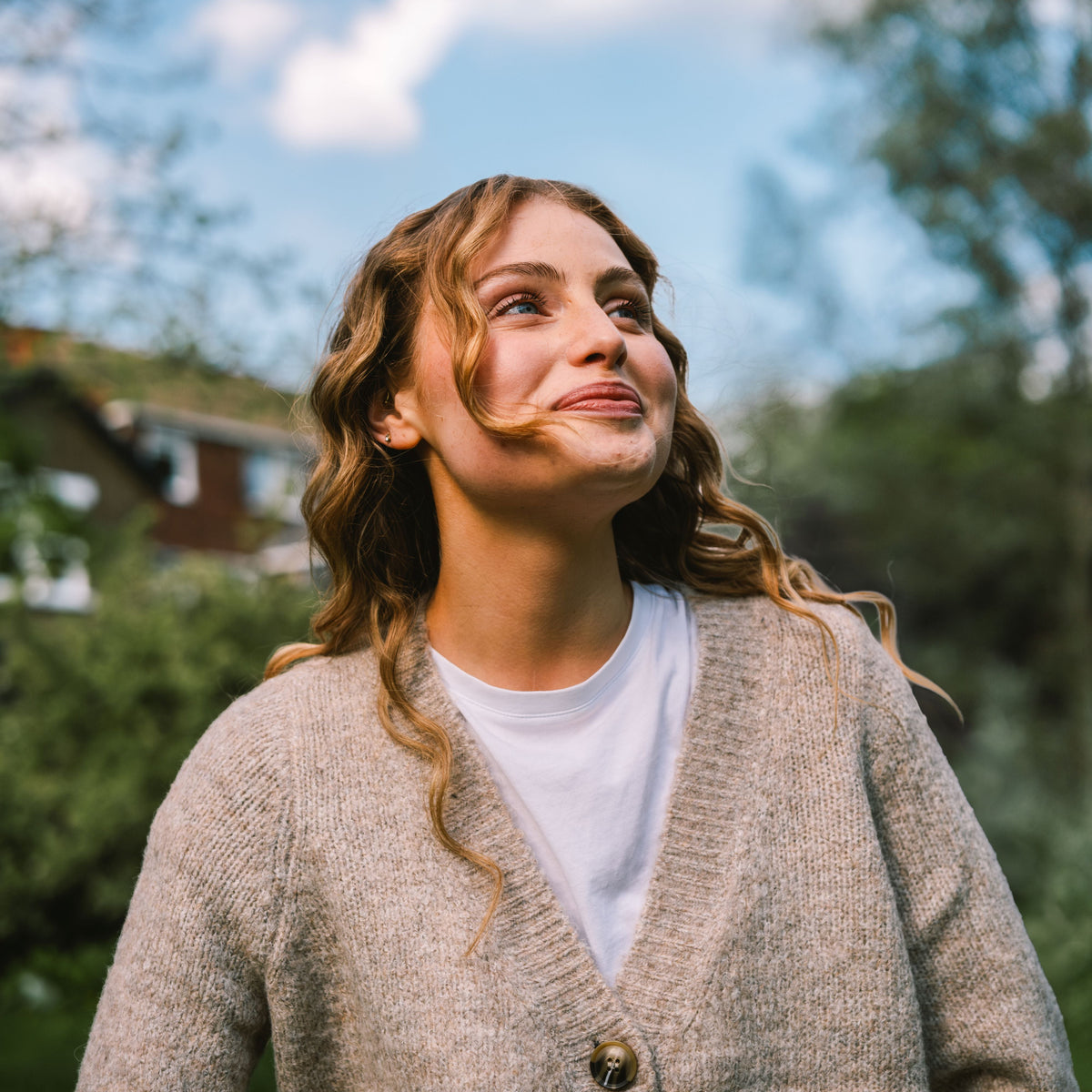 A woman outdoors wearing a beige knit cardigan over a white t-shirt, looking upward against a blurred background of trees and blue sky.